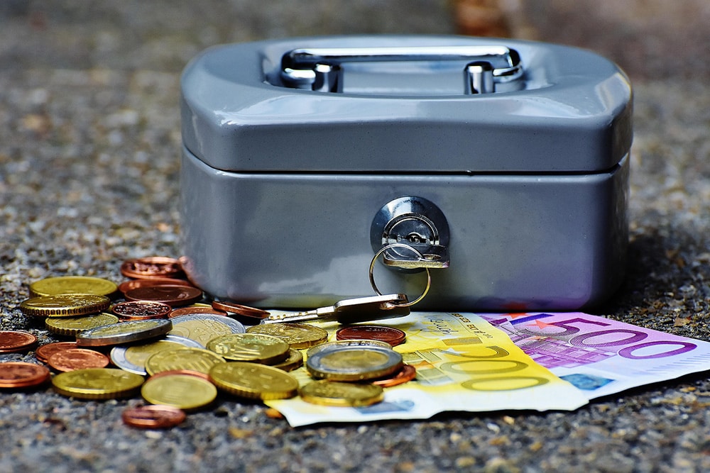 Locked money box on the ground with coins and notes around it